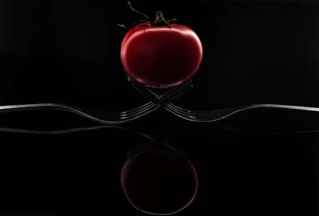 Creative composition of two forks and a red cherry tomato isolated on a black backgroundの写真素材