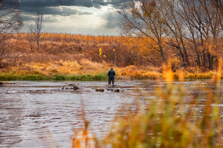 Autumn landscape. A lone fisherman stands knee-deep in the water and fishes against a backdrop of autumn thundery skies and orange autumn foliageの写真素材
