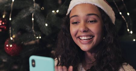 Beautiful Smiling Girl in Santas Hat sitting near Decorated Christmas Tree, Having Video Call via...