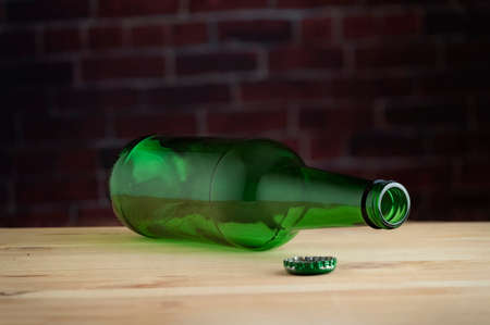 An empty glass beer bottle on a wooden table, against a brick wall.