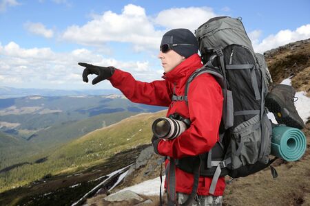 traveler in red jacket with backpack and camera in hand points into the distance hand being on the hillsideの写真素材