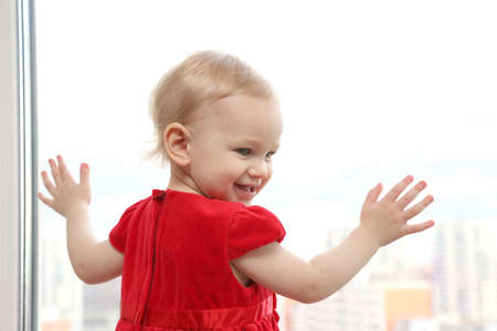 blonde little girl in a red dress smiling next to the windowの写真素材