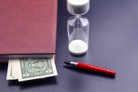 Hourglass, money, pen and notebook are on the table. Business office items. Time is money. Business...