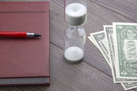 Hourglass, money, pen and notebook are on the table. Business office items. Time is money. Business...