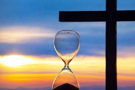 Hourglass has finished counting time on the background of the sunset and a wooden cross. End of...