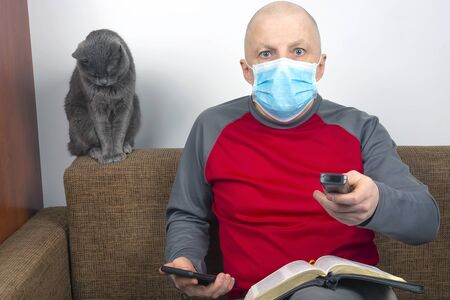 man in quarantine at home with a medical mask on his face lies on the couch and watches TV next to a gray cat. Rest during the coronavirus epidemic.の写真素材