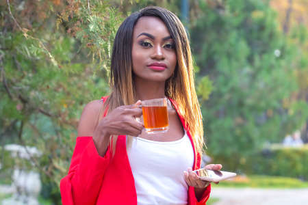 Beautiful young African girl with a Cup of tea and a phone in her hands in the Park