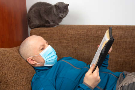 man in a medical mask at home in quarantine because of a coronavirus epidemic reads a bible sitting...