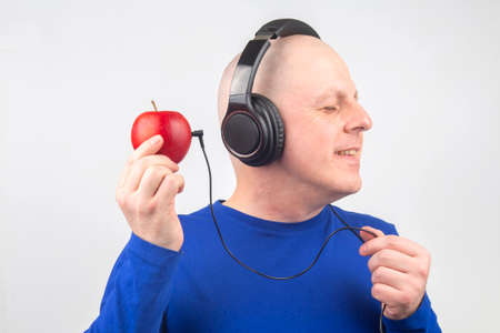 bald man with headphones listens to music through a red apple player. Metaphor and concept of vitamin benefits in music and soundの写真素材