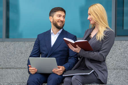 successful businessmen man and woman are sitting on the stairs of a business building with documents and a laptop in their hands discussing business projectsの写真素材