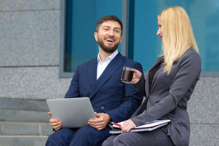 businessmen man and woman sitting on the stairs of a business building with documents and a laptop in their hands drinking coffee and discussing business work planの写真素材