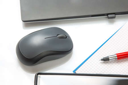 Computer wireless mouse, pen and notepad next to a laptop on a white table close-up. Items for...