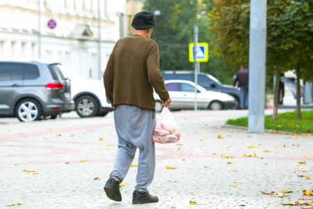 homeless poor man walks down the city street with a bag in his hand. social problems of the city and peopleの写真素材