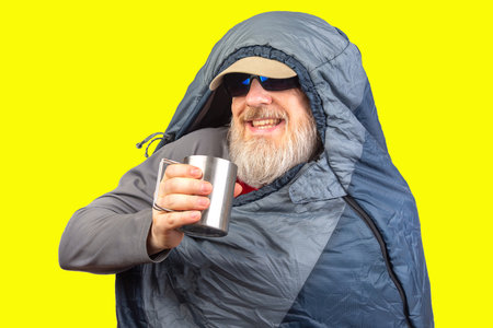bearded man in a sleeping bag with a cup of coffee. equipment for recreation in tourism and travel