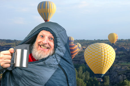 Happy bearded man in a sleeping bag with a cup of coffee against the backdrop of nature in the mountains. nature hikes in the mountainsの写真素材