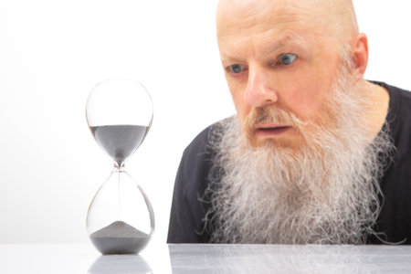 A contemplative man with a long white beard intently observes an hourglass, reflecting on the passage of time. The minimalist white background highlights the stark contrast between the man's expression and the flowing sandの写真素材