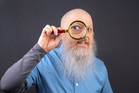 Serious Bearded Man Using Magnifying Glass for Close Examination. One Eye Magnified. search and research concept. detective agencyの写真素材