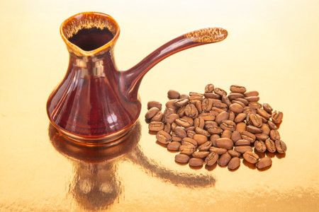 Traditional red ceramic cezve with a speckled amid handlest a pile of coffee beans on a shiny gold surface, ideal for gourmet coffee and kitchen decor themesの写真素材