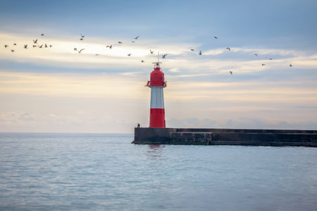 A serene coastal scene with a red and white lighthouse on a pier, a calm sea under a soft pastel sky at dawn.の写真素材