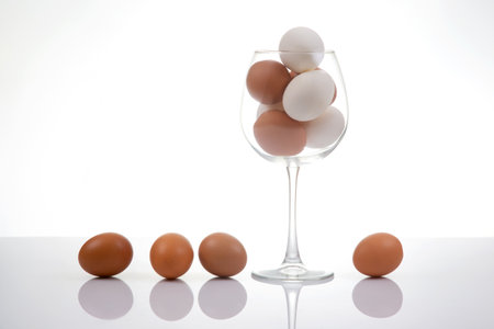 White and brown eggs stacked in a clear wine glass, accompanied by three eggs on a glossy white background, offering a clean and modern food photography sceneの写真素材