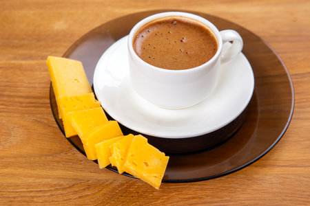 An overhead shot of a frothy espresso in a white cup, resting on a saucer, accompanied by cheese slices on a cozy wooden table surfaceの写真素材