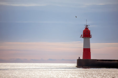 Red and white lighthouse stands tall on a pier, set against a calm sea and a soft, cloudy sky during a peaceful morning.の写真素材