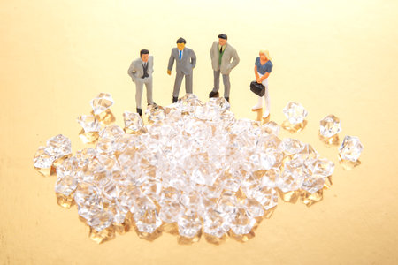 Miniature business people standing around a pile of shiny crystals on a golden background, symbolizing wealth and success in a corporate setting.の写真素材