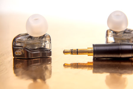 Elegant earbud with a white tip, a 3.5mm plug, and a black music player on a shiny gold backdrop,...