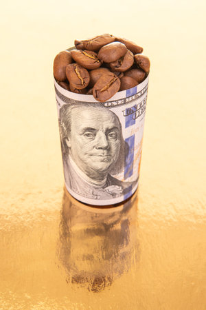 On a reflective gold surface, a rolled dollar bill filled with coffee beans standing beside scattered beans, illustrating the concept of trade value.の写真素材