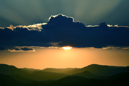 Scenic sunset with the sun peeking through a dark cloud. The sky is bathed in warm golden hues, while the illuminated cloud edges add depth and contrast to the image.の写真素材