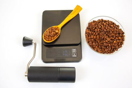 Close-up of a black coffee grinder, a digital scale holding a wooden spoon with roasted beans, and a glass bowl of coffee beans on a clean white surface for brewing visualsの写真素材