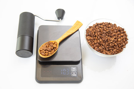 A black manual coffee grinder, a digital scale with a wooden spoon of roasted coffee beans, and a glass bowl of beans on a white background, ideal for coffee preparationの写真素材