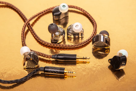 Close-up of three earbuds with woven cables and gold-plated jacks on a glossy golden background,...