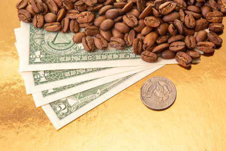 US dollar bills are partially covered by roasted coffee beans on a golden background, highlighting...