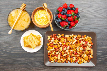 A wooden table displays a healthy spread: a bowl of honey with a dipper, a bowl of butter, a plate of honeycomb, a mix of nuts on a tray, and a bowl of fresh strawberries and blueberries.の写真素材
