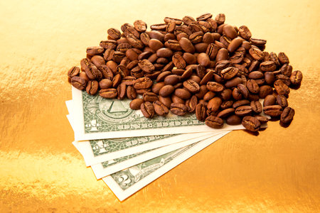 Roasted coffee beans cascade over a stack of US dollar bills on a golden backdrop, representing the fusion of coffee trade and financial wealth in a luxurious setting.の写真素材