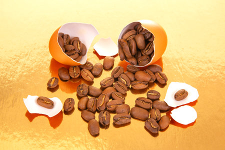 Creative still life of coffee beans emerging from a broken egg on a gold background, with eggshell...
