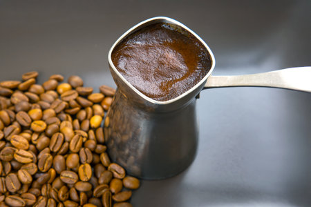 Close-up of a silver cezve with frothy Turkish coffee on a gray surface, surrounded by roasted coffee beans. Ideal for coffee culture, beverage ads, or rustic decor.の写真素材