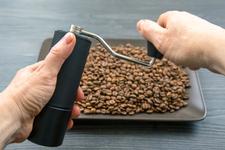 Hands operating a black manual coffee grinder over a tray of roasted coffee beans on a dark wooden surface, capturing the coffee grinding processの写真素材