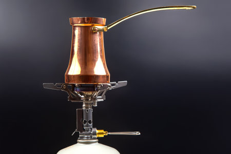A shiny copper Turkish coffee pot with a brass handle sits atop a portable gas stove with a white fuel canister, set against a sleek dark background, perfect for coffee brewingの写真素材