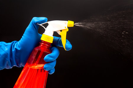 A hand wearing a blue glove sprays a red cleaning bottle with a yellow nozzle, creating a mist...