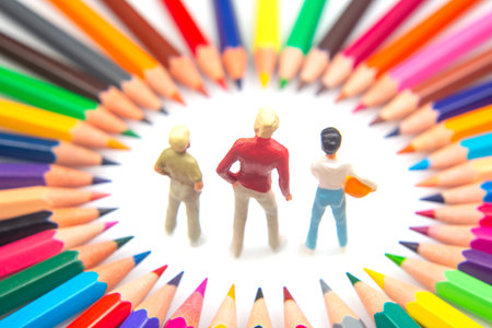 Three figurines are encircled by a spectrum of colored pencils, arranged in a starburst pattern, highlighting creativity and imagination on a white surfaceの写真素材