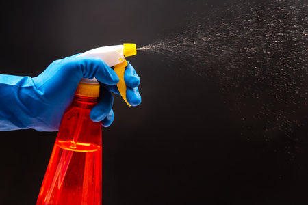 A close-up of a hand in a blue glove spraying a red cleaning bottle with a yellow trigger, mist dispersing against a dark background, ideal for hygiene or cleaning conceptsの写真素材