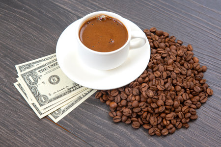 White cup of freshly brewed coffee on a saucer, surrounded by roasted coffee beans and dollar bills, placed on a dark wooden surface.の写真素材