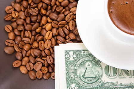 Aromatic morning coffee in a white cup, served on a saucer with a pile of coffee beans and dollar bills, reflecting the value of premium coffee.の写真素材