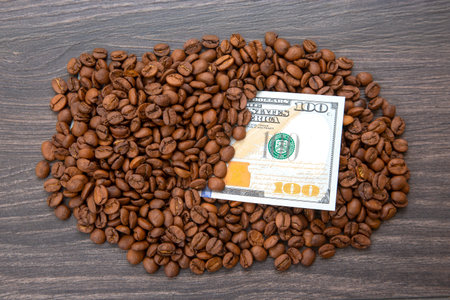 hundred-dollar bill partially covered by roasted coffee beans on a dark wooden surface. Concept of...