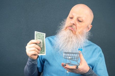 bald man with a large white beard holds a dollar and a model of a grocery basket in his hands....
