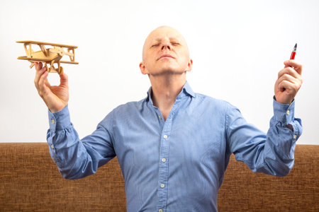 Dream of flight. inventor concept. man with a bald head and a blue shirt joyfully raises a wooden airplane model in one hand and a red pen in the other, expressing delight in a relaxed indoor space.の写真素材