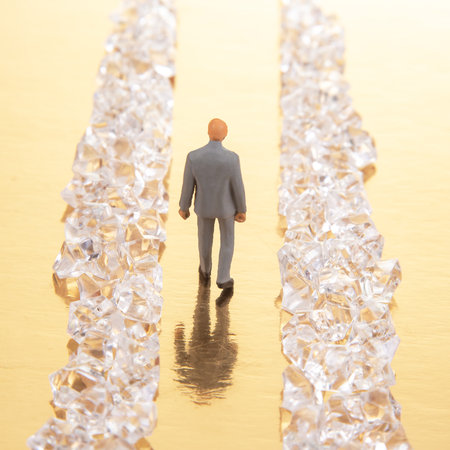 A miniature businessman in a gray suit walks confidently between two rows of sparkling crystal formations on a reflective golden surface, symbolizing success and wealth.の写真素材