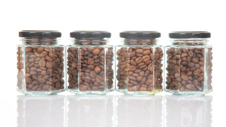 Four glass jars with black lids filled with roasted coffee beans, arranged in a row on a reflective surface against a white backgroundの写真素材
